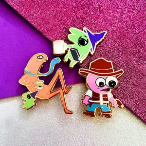 Peut inclure: Trois épingles en émail représentant des personnages de dessins animés. L'une représente un personnage rose avec un chapeau et un gilet de cow-boy, une autre un personnage vert avec un livre, et la dernière un personnage orange avec une tasse.