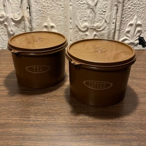 Può includere: Due contenitori di plastica marroni con coperchi. Il contenitore a sinistra dice "TEA" e il contenitore a destra dice "COFFEE".