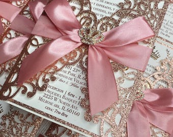 May include: Rose gold glitter laser cut invitation with a pink satin bow and a crown design. The invitation text reads "101 23rd Ave, Rose Park, IL 60160, Adrinos de Velacio, Lover & Allan".