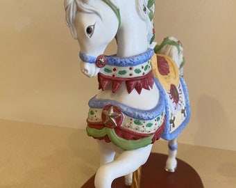 Può includere: Una statuetta di cavallo di carosello in porcellana bianca con una sella colorata e nastri blu e verdi. Il cavallo è in piedi su una base di legno.