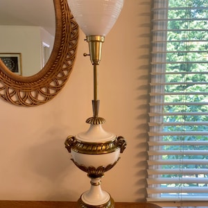 May include: A white and gold floor lamp with a white, textured glass shade. The lamp base is shaped like a vase with gold accents and lion head handles.