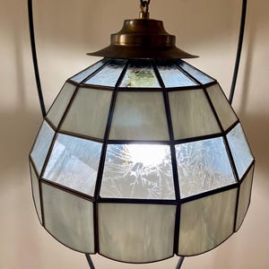 May include: A white stained glass pendant light fixture with a brass ceiling mount and a black metal stand. The light fixture has a geometric design with a frosted glass finish.