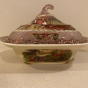 May include: A brown and white ceramic serving dish with a lid. The lid has a raised handle and features a floral design. The dish has a similar floral design and a scalloped edge.