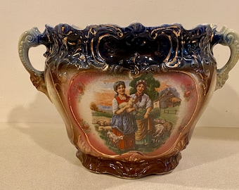 May include: A large, ornate, blue and gold ceramic planter with a scene of two women and sheep in a field. The planter has two handles and a scalloped rim.