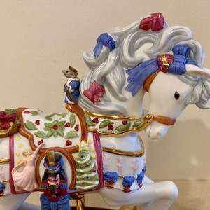 May include: A white porcelain rocking horse with a gold trim and a Christmas theme. The horse is decorated with holly, bows, and a nutcracker. A ballerina and a mouse are also on the horse.
