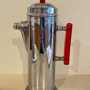 May include: A vintage silver cocktail shaker with a red plastic top and handle. The shaker has a spout on the side and a rounded base.
