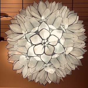 May include: A white, flower-shaped pendant light fixture with a layered petal design and black trim. The light is hanging from a dark metal grid, creating a decorative and functional lighting element.