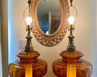 May include: Two amber glass table lamps with ornate brass bases and accents. The lamps have a bulbous shape and are topped with a simple metal arm holding a lightbulb. A woven oval mirror hangs in the background.