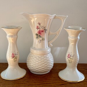 May include: A set of three white ceramic vases with a floral design. The vases have a textured surface and gold accents. The set includes two candle holders and one pitcher.