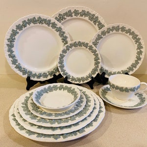 May include: A set of white ceramic dinnerware with a green leafy pattern. The set includes a teacup and saucer, a bowl, and five plates of varying sizes.