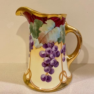 May include: A vintage porcelain pitcher with a gold rim and handle. The pitcher is decorated with a hand-painted design of purple grapes and green leaves on a cream background.