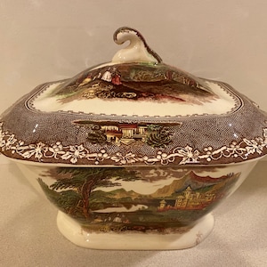 May include: A brown and white ceramic serving dish with a lid. The dish features a floral pattern and a landscape scene with a building and trees.