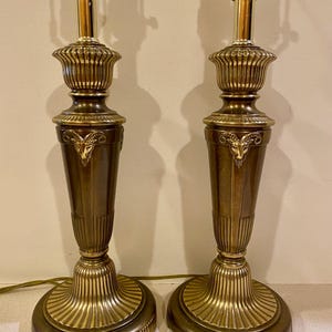 May include: Pair of brass table lamps with a classic design. Each lamp features a fluted base, a detailed central section with a ram's head, and a decorative top. The lamps have a warm, golden hue and are designed for indoor use.