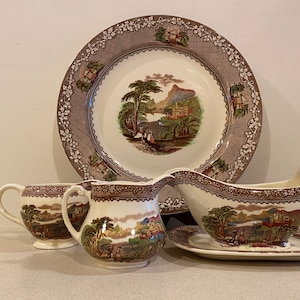 May include: A set of brown and white vintage china featuring a landscape scene. The set includes a large dinner plate, two creamer pitchers, a gravy boat, and a saucer.