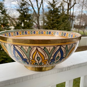 May include: A large, ceramic bowl with a gold rim and intricate blue, green, and yellow floral patterns. The bowl is sitting on a white wooden railing.