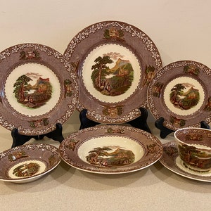 May include: A set of brown and white ceramic dinnerware with a scenic landscape design. The set includes a large dinner plate, a salad plate, a soup bowl, a teacup, and a saucer.