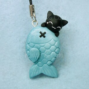 Puede incluir: Una figura de gato negro posada sobre un dije de pez azul claro. El pez tiene un patrón de escamas y una cruz negra. El dije está unido a un cordón negro con un cierre plateado. El texto "Kawai & Cute" es visible.