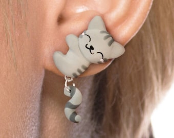 May include: Gray and white cat-shaped dangle earrings with a smiling face. The cat's tail dangles from the bottom of the earring.
