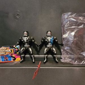 May include: Two black and silver action figures with skeletal faces and blue accents. The figures are posed in a fighting stance. The figures are from a line of wrestling action figures called "Heroes de la Lucha Libre".