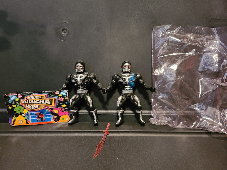 May include: Two black and silver action figures with skeletal faces and blue accents. The figures are posed in a fighting stance. The figures are from a line of wrestling action figures called "Heroes de la Lucha Libre".