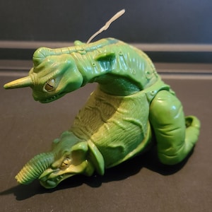 May include: A green plastic toy dragon with two heads. The dragon is curled up in a defensive position with its heads facing forward.