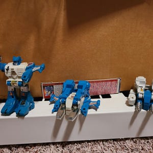 May include: Three vintage Transformers action figures in blue, white, and silver. One figure is in robot mode, holding a silver weapon. Another is in vehicle mode, and the third is in a different vehicle mode.