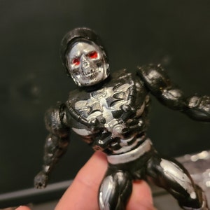 May include: A black and silver action figure with a skull head and red eyes. The figure is wearing a black and silver suit with a skeletal design.