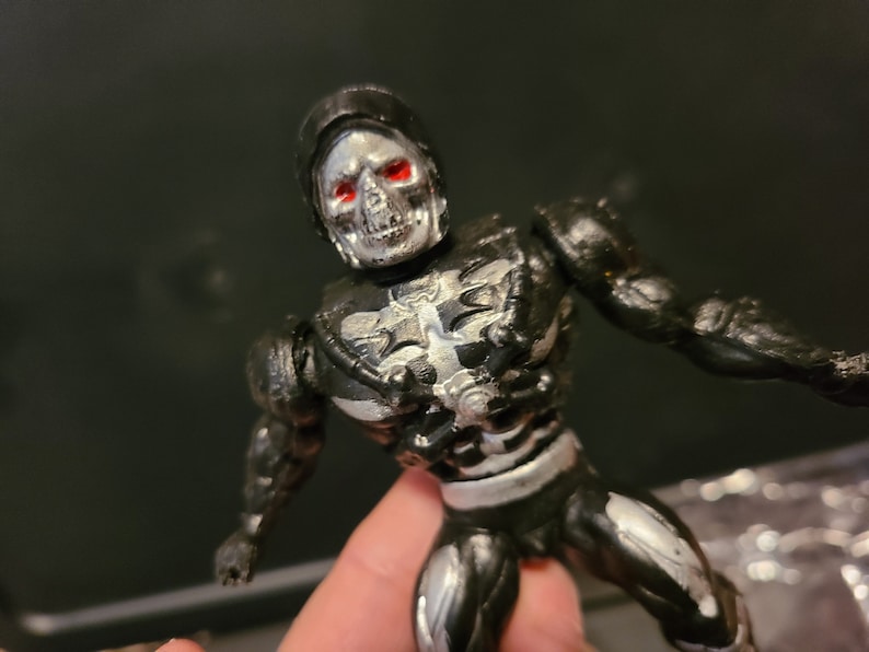 May include: A black and silver action figure with a skull head and red eyes. The figure is wearing a black and silver suit with a skeletal design.