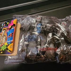 May include: Two silver and black action figures of wrestlers from the "Heroes de la Lucha Libre" brand. The figures are packaged in a clear plastic bag with a colorful card featuring the brand logo and wrestlers.