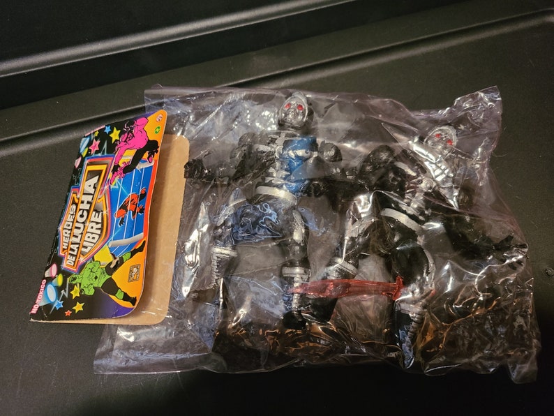 May include: Two silver and black action figures of wrestlers from the "Heroes de la Lucha Libre" brand. The figures are packaged in a clear plastic bag with a colorful card featuring the brand logo and wrestlers.