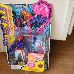 May include: Action figure packaging featuring a red-haired action figure in a blue and white suit with a red belt. The figure is holding a gun. The packaging is blue, yellow, and pink with the words "SPARTAN" and "STREET FIGHTERS" printed on it.