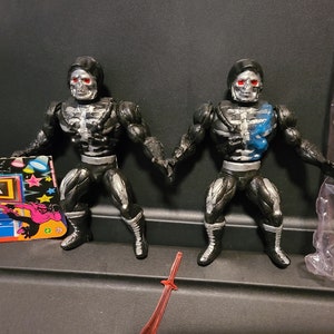 May include: Two black and silver skeleton action figures with red eyes and black cloaks. The figures are posed in a fighting stance. One figure has a blue paint splatter on its chest.