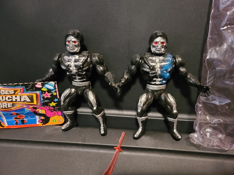 May include: Two black and silver skeleton action figures with red eyes and black cloaks. The figures are posed in a fighting stance. One figure has a blue paint splatter on its chest.