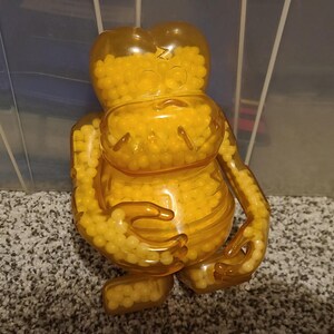 May include: A yellow plastic figurine shaped like a person filled with small yellow beads. The figurine has a rounded head and a wide, smiling mouth.