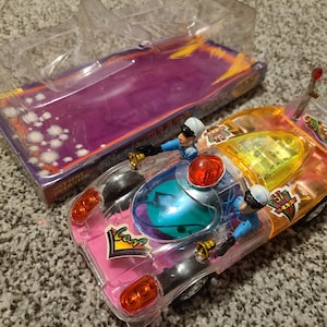 May include: A clear plastic toy car with a pink, yellow, and blue design. The car has two figures inside, a police officer and a driver. The car has a light and a siren on top. The car has the words "Zoom" and "51" on the side.