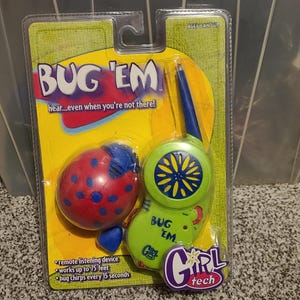 May include: A packaged "Bug 'Em" remote listening device toy. The packaging is yellow and green, with a ladybug-shaped receiver and a green transmitter. The text "BUG 'EM" is prominently displayed, along with "Girl Tech" branding.