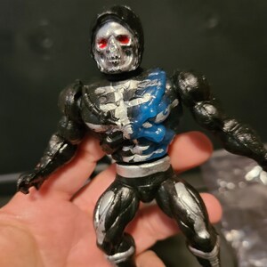 May include: Black and silver skeleton action figure with blue accents. The figure has a skull head with red eyes and is wearing a black hooded cape.