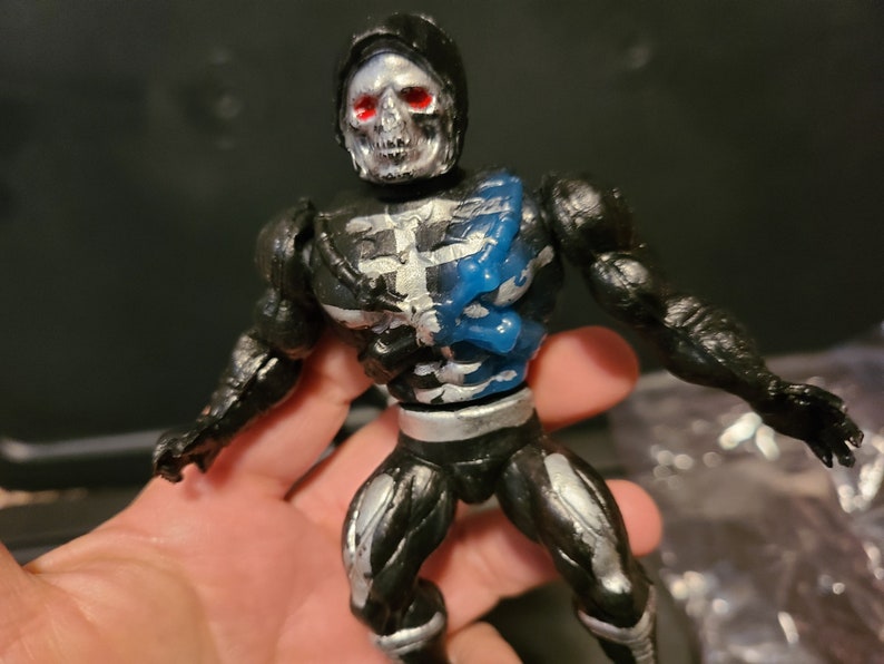 May include: Black and silver skeleton action figure with blue accents. The figure has a skull head with red eyes and is wearing a black hooded cape.