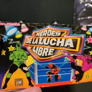 May include: A colorful card with the words "H&Eacute;ROES DE LA LUCHA LIBRE" in a yellow circle. The card features three wrestlers in a wrestling ring. The wrestlers are wearing green, pink, and red costumes. The card also has the words "NO T&Oacute;XICO" and "HECHO EN M&Eacute;XICO" printed on it.