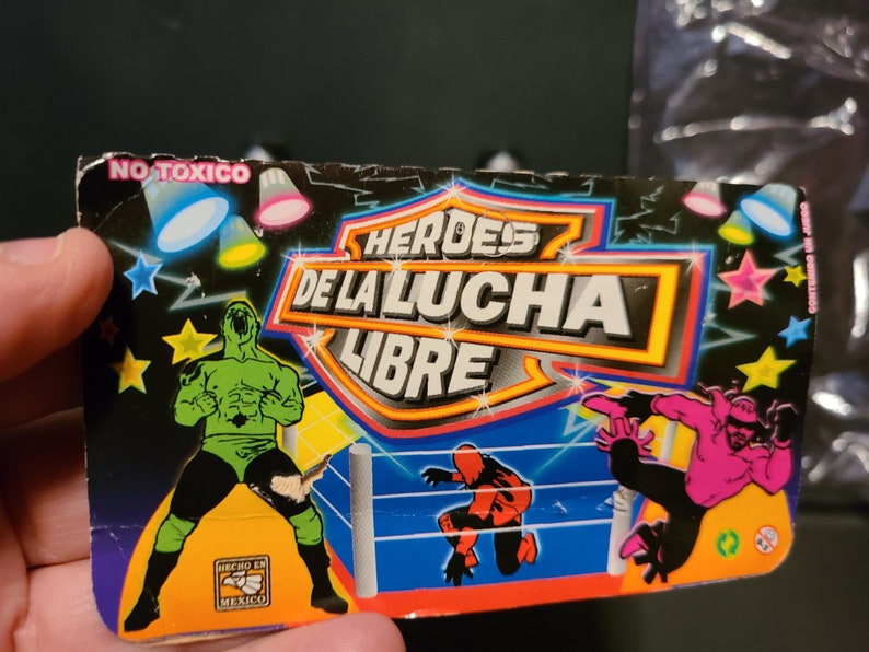 May include: A colorful card with the words "H&Eacute;ROES DE LA LUCHA LIBRE" in a yellow circle. The card features three wrestlers in a wrestling ring. The wrestlers are wearing green, pink, and red costumes. The card also has the words "NO T&Oacute;XICO" and "HECHO EN M&Eacute;XICO" printed on it.
