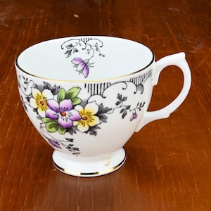 Op de afbeelding: Witte theekop met een gouden rand en oor, versierd met een bloemmotief van paarse, gele en witte bloemen. De kop heeft een voetstuk en staat op een houten oppervlak.