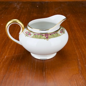 Puede incluir: Cremera de porcelana blanca con asa y ribetes dorados. La cremera presenta una banda decorativa con motivos florales rosas, verdes y morados. Tiene un cuerpo redondeado y un pico vertedor.