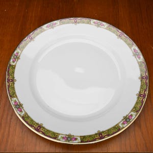 Puede incluir: Un plato de porcelana blanco con un borde decorativo. El borde presenta un diseño verde y morado con detalles florales rosas y ribete dorado. El plato es redondo y tiene una superficie lisa.