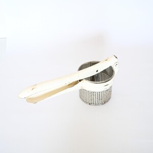 May include: White metal potato masher with a long handle and a perforated cylinder. The masher is designed for crushing cooked potatoes.