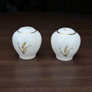 May include: A pair of white ceramic salt and pepper shakers with a gold wheat stalk design. Each shaker has a gold band around the top and small holes for dispensing. The shakers are on a dark brown surface.