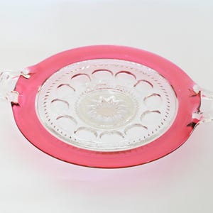 Puede incluir: Un plato de servir de vidrio transparente con un borde rosa y dos asas. El plato tiene un pozo central y un anillo de pequeñas hendiduras alrededor del borde.