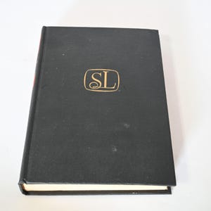 Puede incluir: Libro de tapa dura negro con letras doradas en el lomo y un logotipo en relieve dorado en la portada que dice "SL".