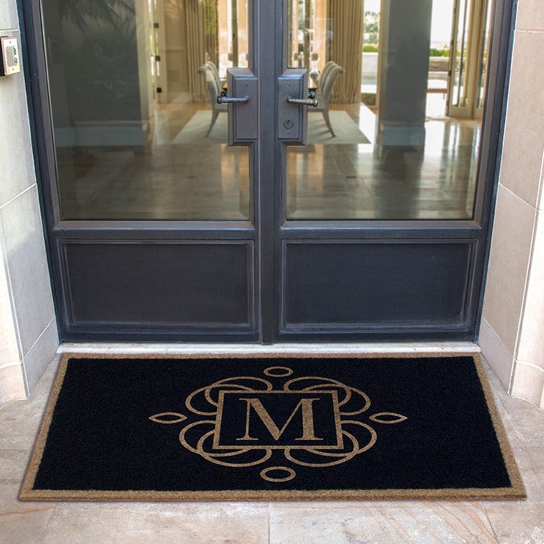 May include: A black doormat with a brown border and a monogram of the letter "M" in a decorative frame.