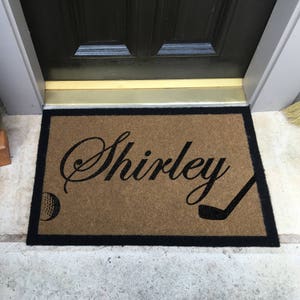 以下が含まれることがあります： 黒い縁取りのある茶色の玄関マットに、筆記体で「Shirley」という名前が書かれています。マットの上には、黒いゴルフクラブと白いゴルフボールも置かれています。