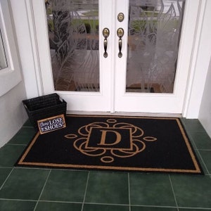 May include: A black doormat with a tan border and the letter "D" in a decorative frame. The doormat is on a green tiled floor. A wooden sign with the text "Please Remove Your Shoes!" is on the floor next to the doormat.